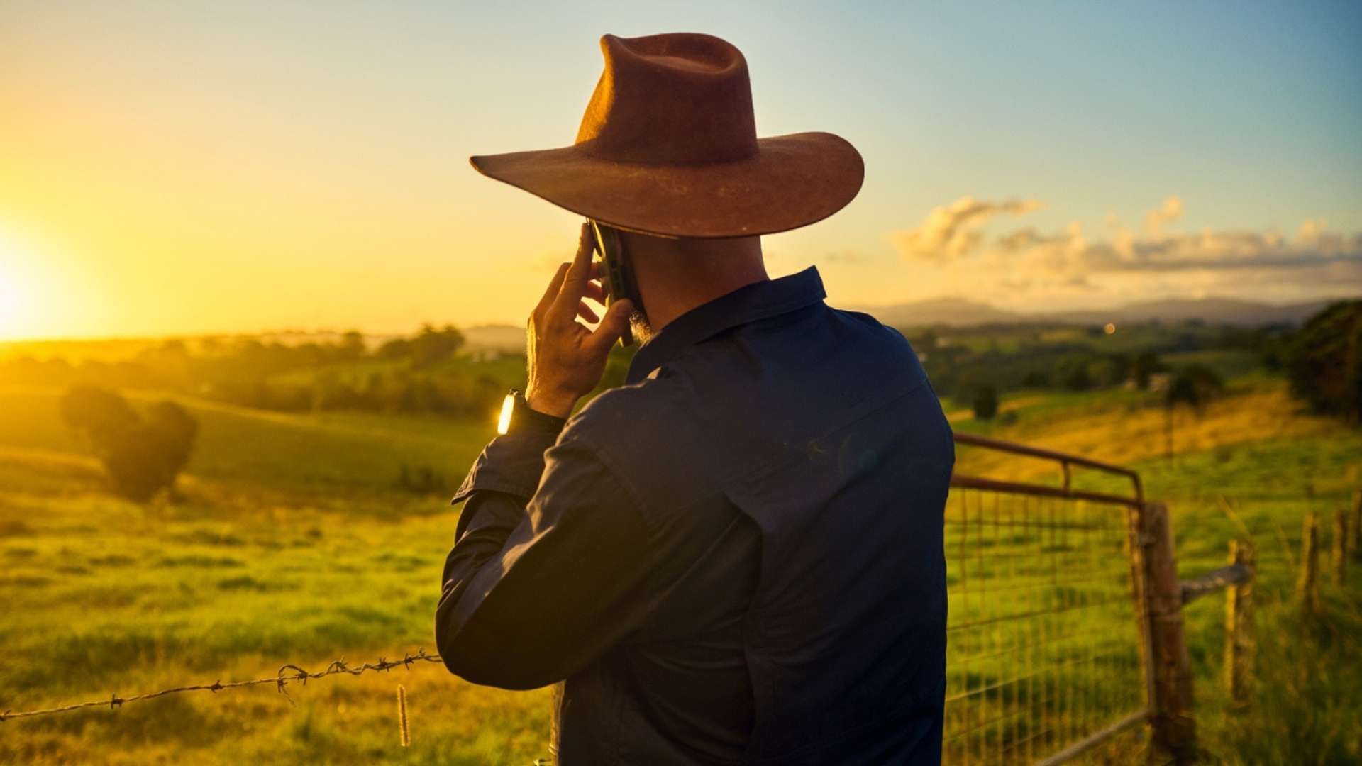 Person standing by a fence in a rural landscape, talking on a smartphone.