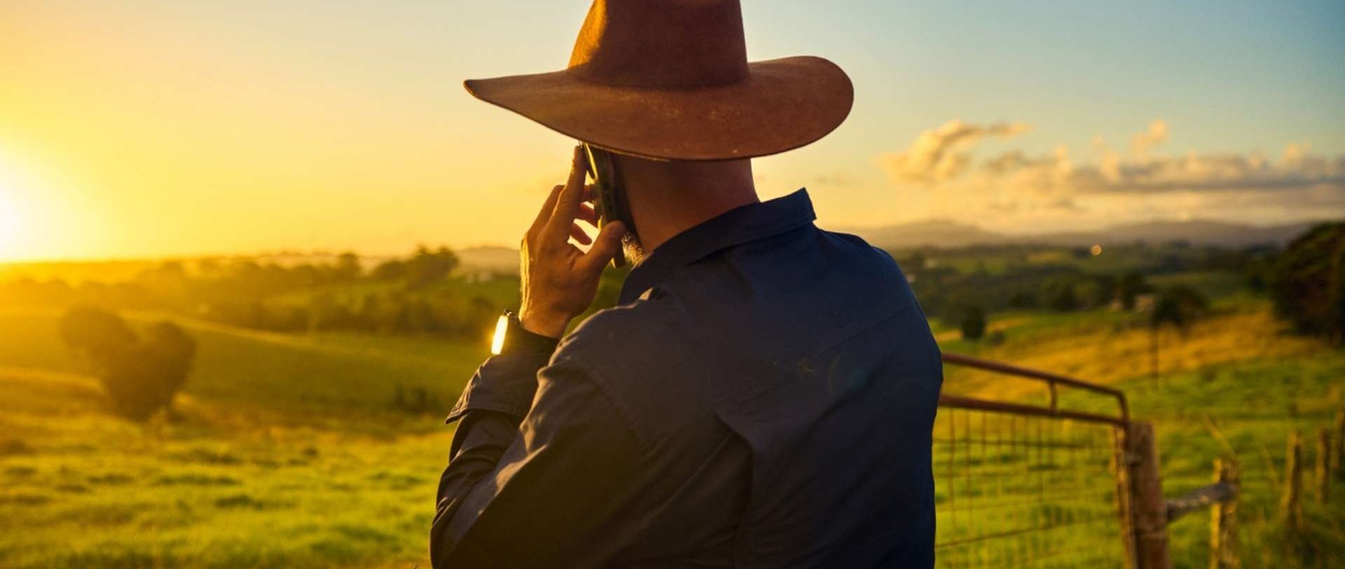 Person standing by a fence in a rural landscape, talking on a smartphone.