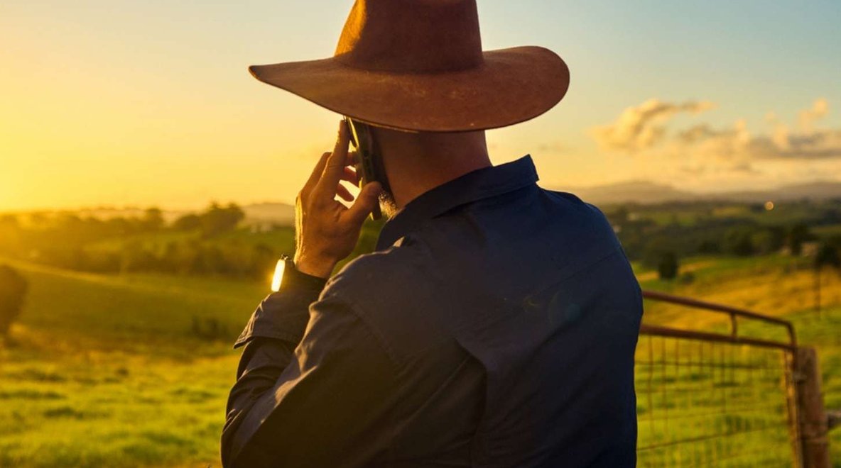 Person standing by a fence in a rural landscape, talking on a smartphone.