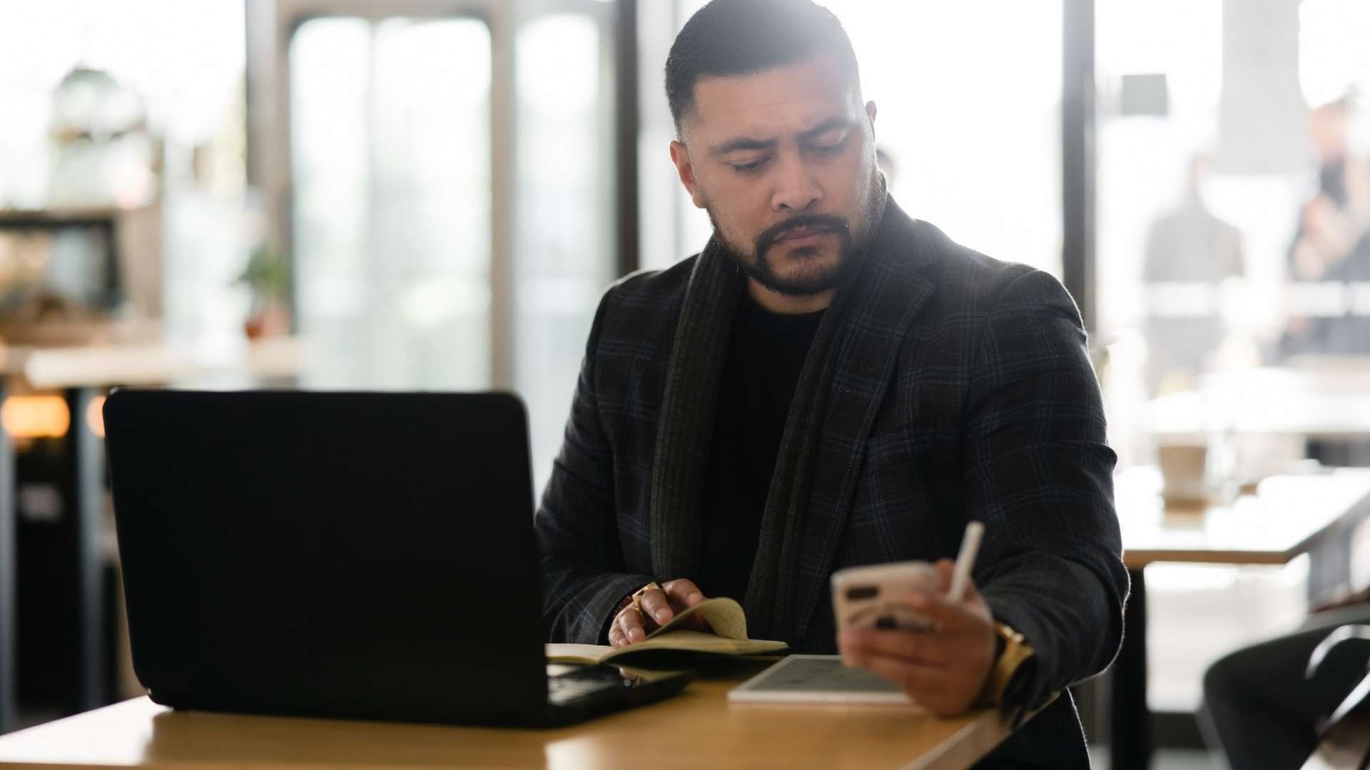 Person using a smartphone and writing in a notebook beside a laptop