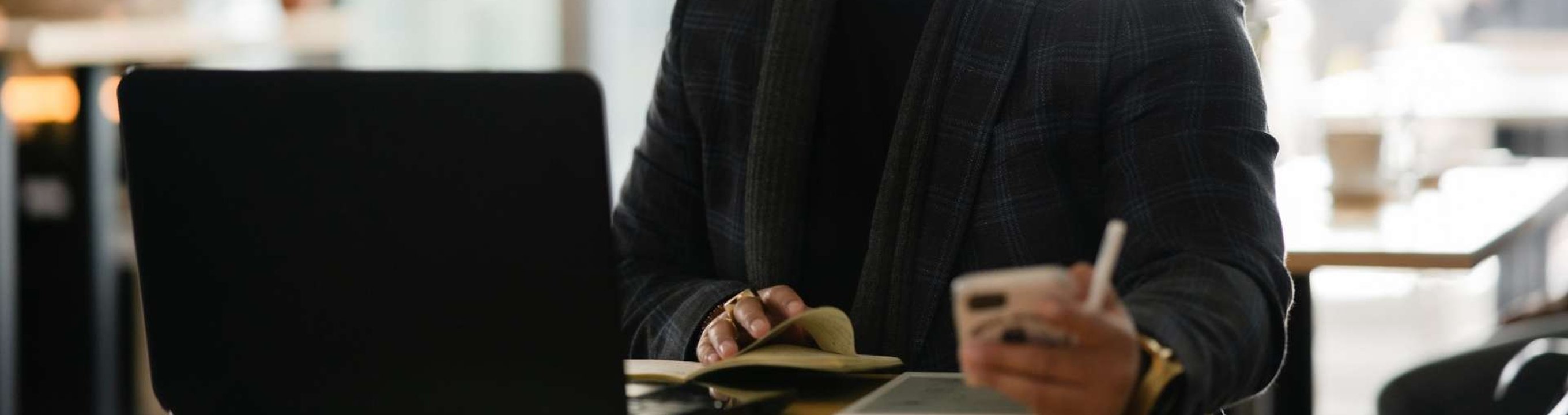 Person using a smartphone and writing in a notebook beside a laptop
