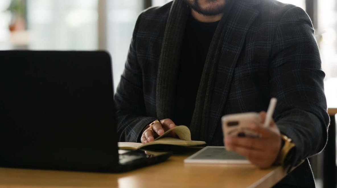 Person using a smartphone and writing in a notebook beside a laptop