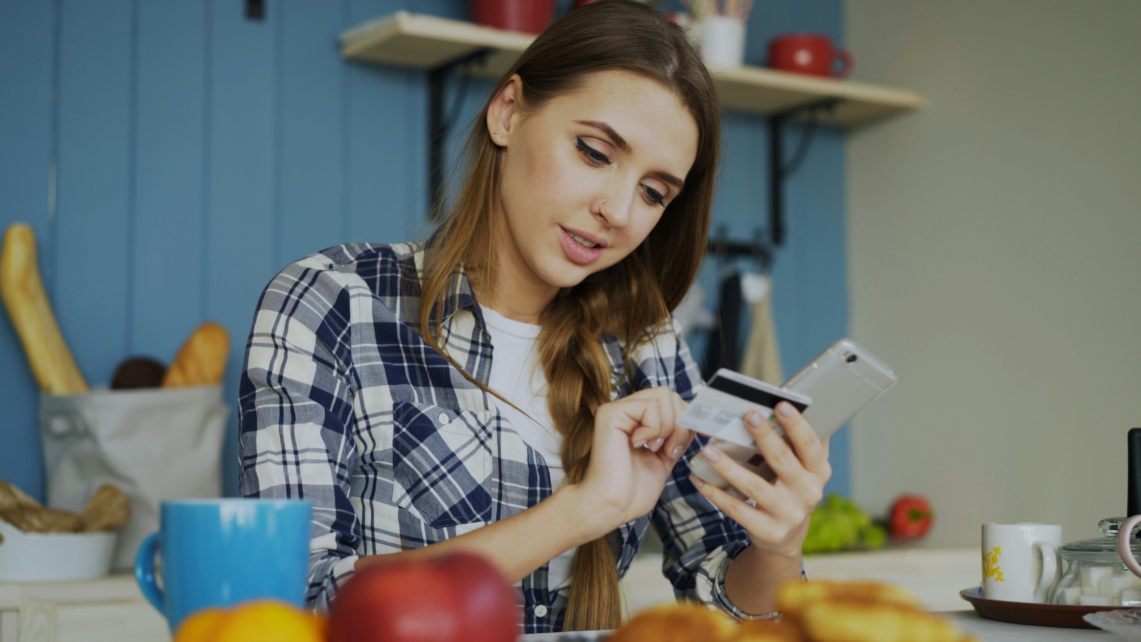 Woman at kitchen table looking at her phone and holding a bank card while making a payment, with food and a mug in the foreground.