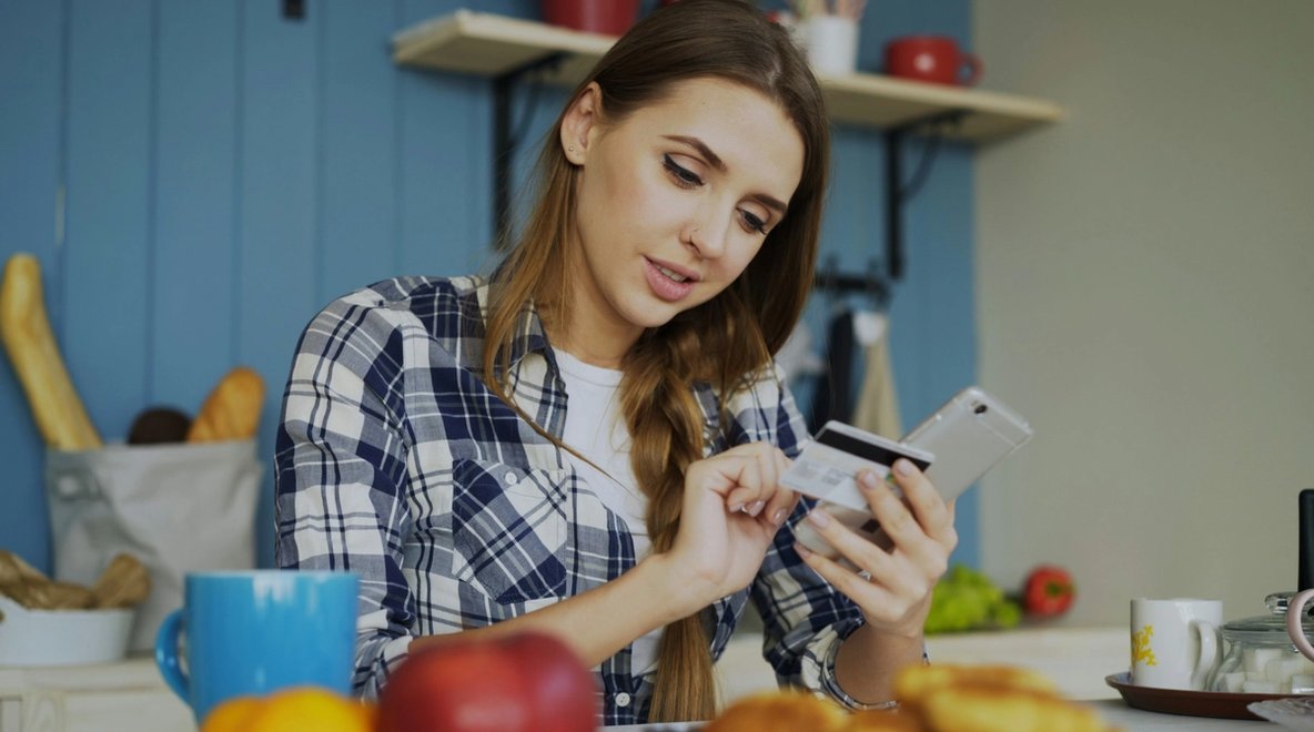 Woman at kitchen table looking at her phone and holding a bank card while making a payment, with food and a mug in the foreground.