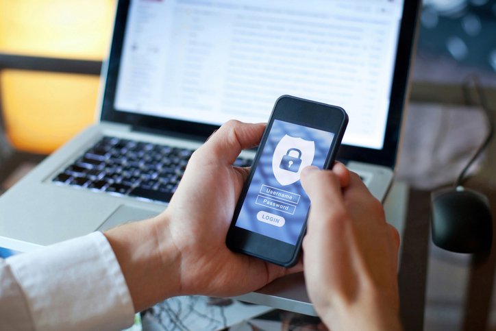 Person holding a smartphone showing a secure login screen with a padlock icon, with a laptop in the background.