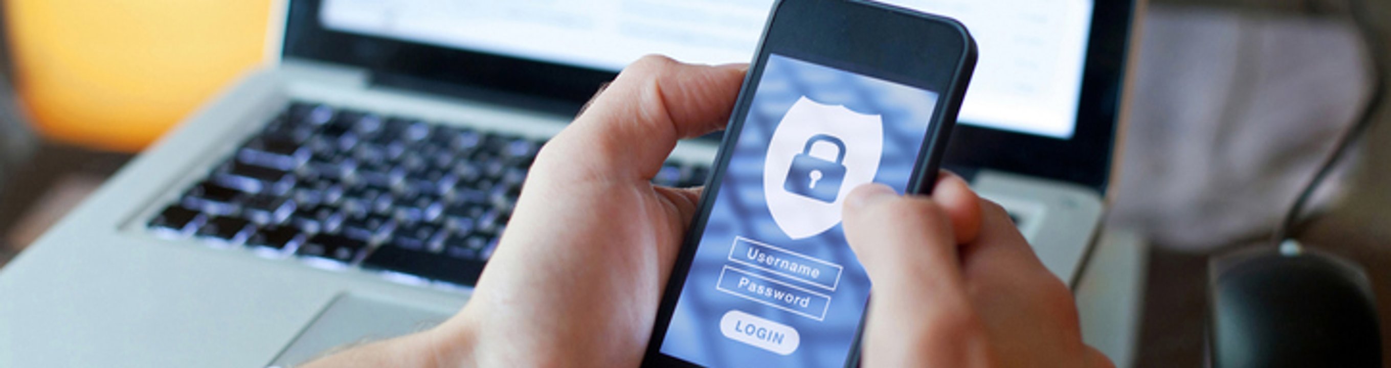 Person holding a smartphone showing a secure login screen with a padlock icon, with a laptop in the background.