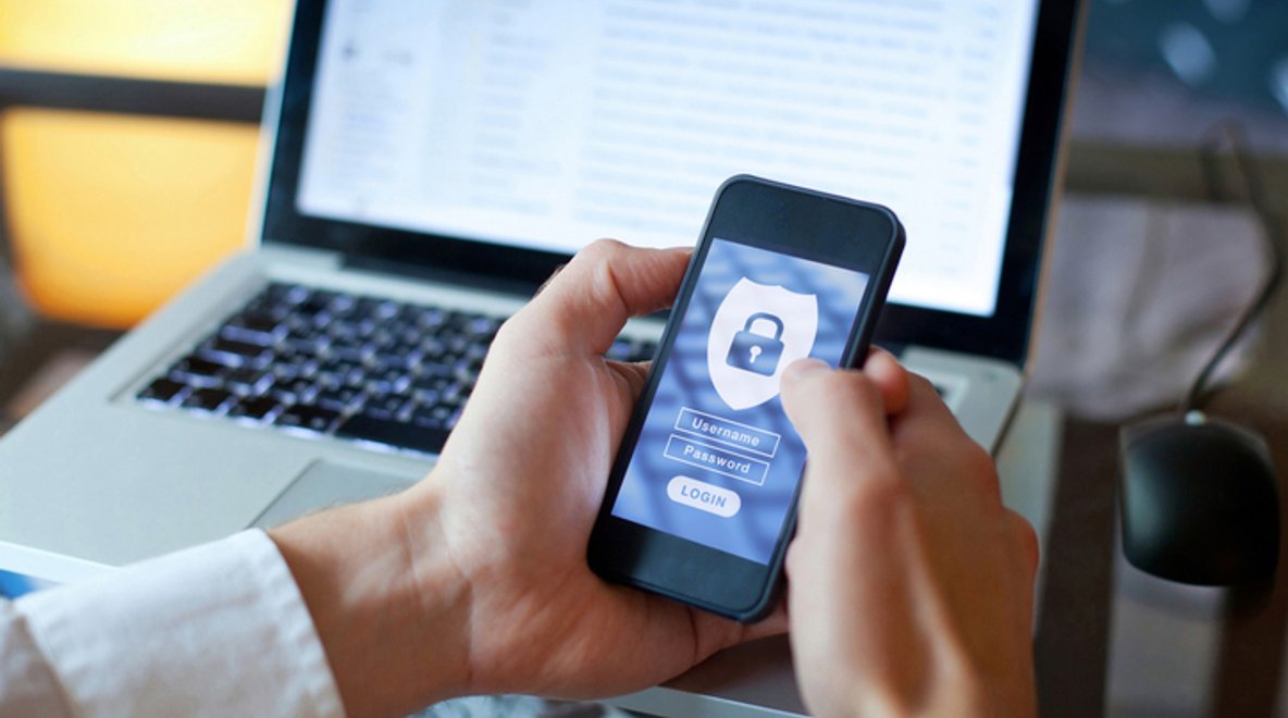 Person holding a smartphone showing a secure login screen with a padlock icon, with a laptop in the background.