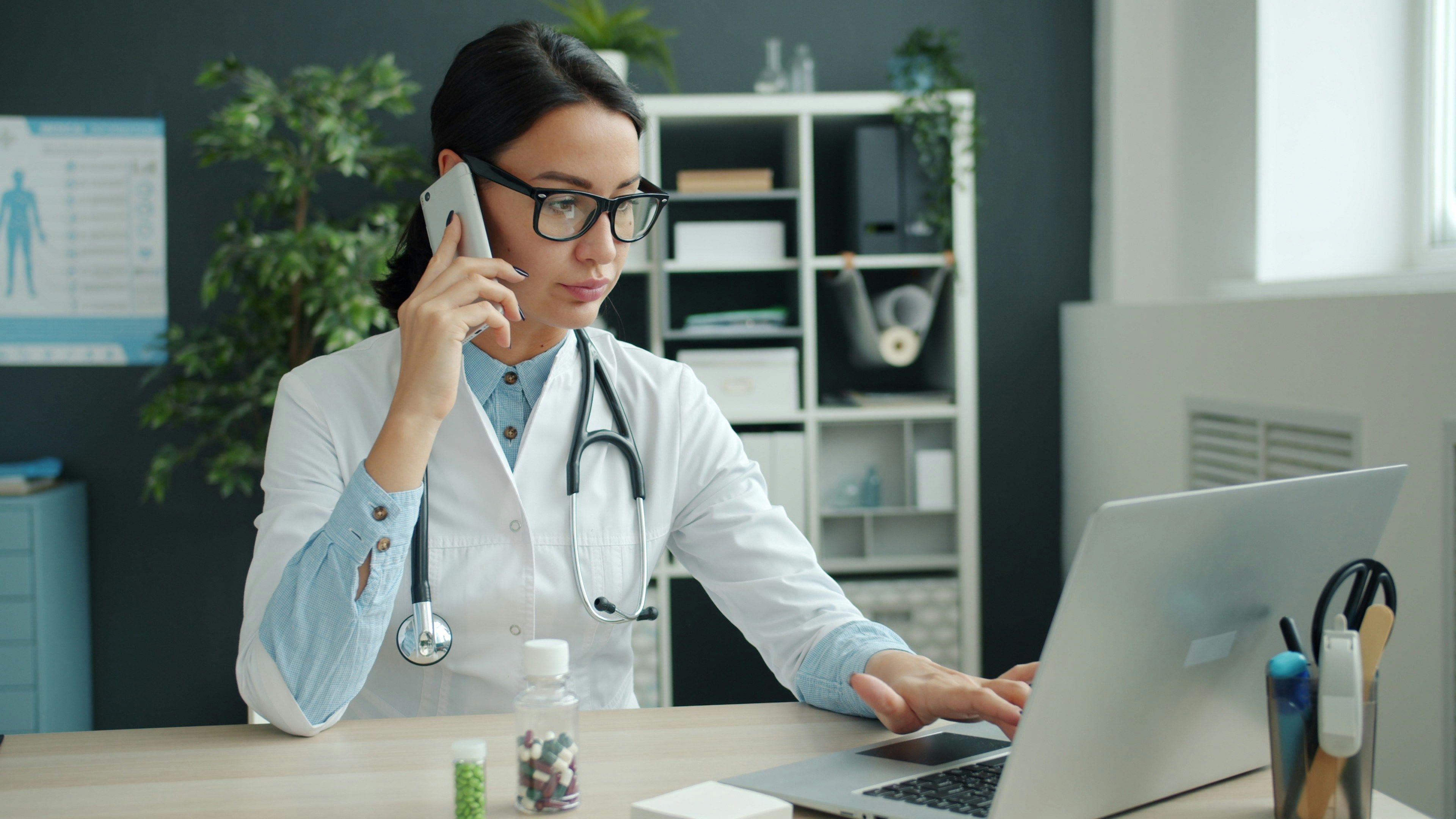 Doctor using a phone while working on a laptop at a desk