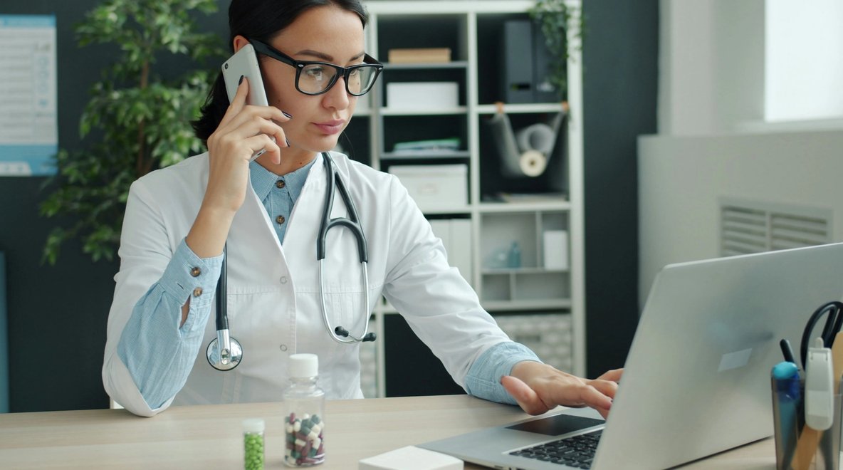 Doctor using a phone while working on a laptop at a desk