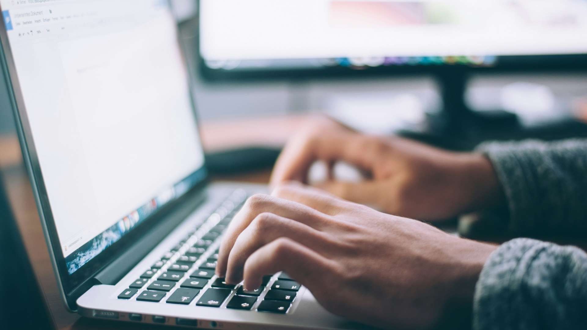 Close-up of hands typing on a laptop keyboard, with a blurred screen and monitor in the background.