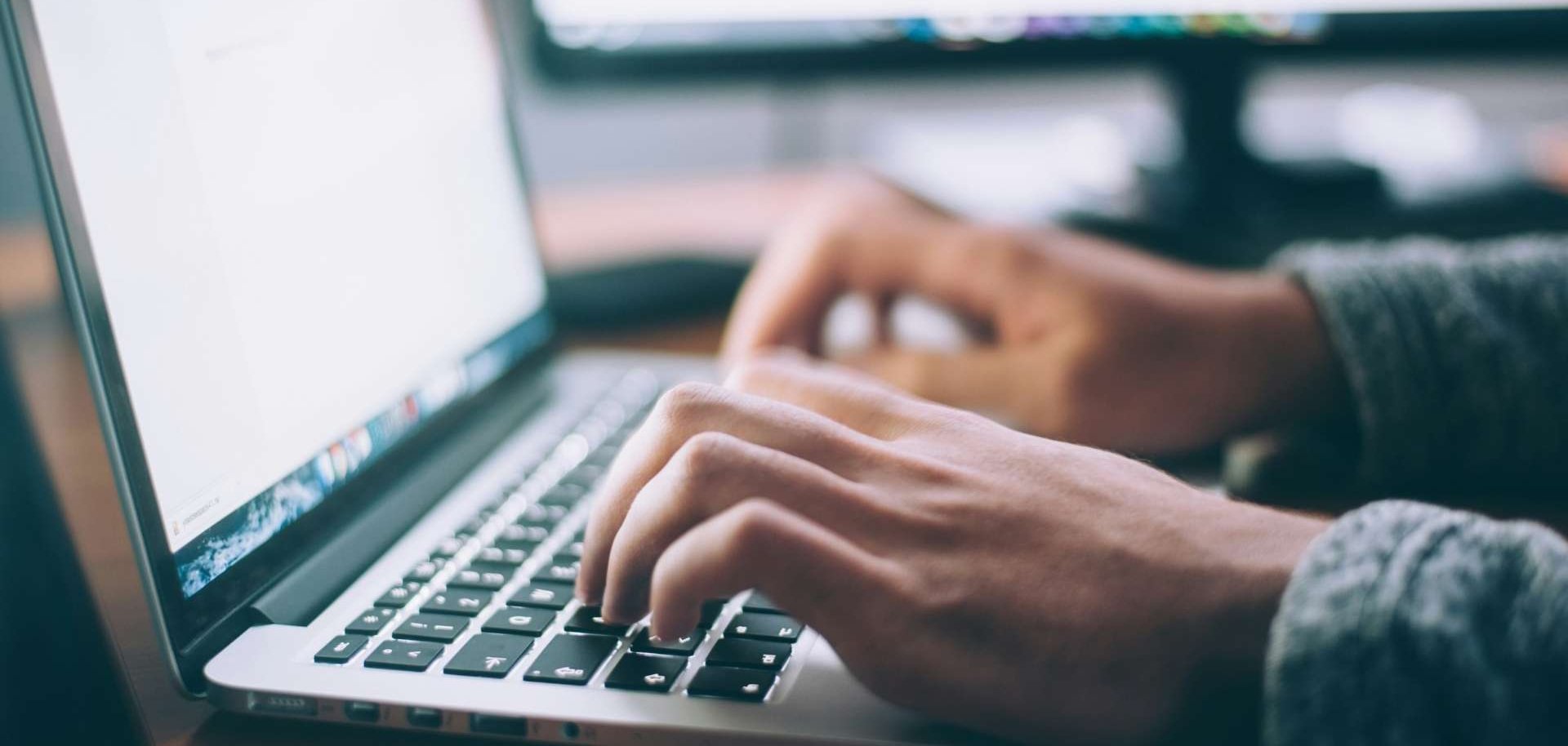 Close-up of hands typing on a laptop keyboard, with a blurred screen and monitor in the background.
