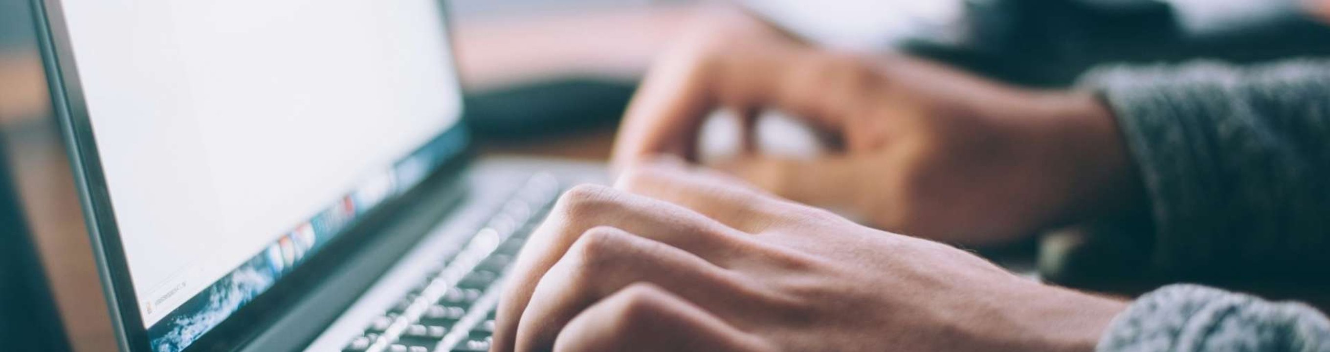Close-up of hands typing on a laptop keyboard, with a blurred screen and monitor in the background.