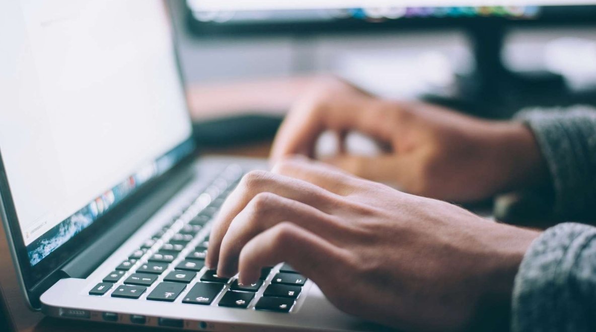Close-up of hands typing on a laptop keyboard, with a blurred screen and monitor in the background.