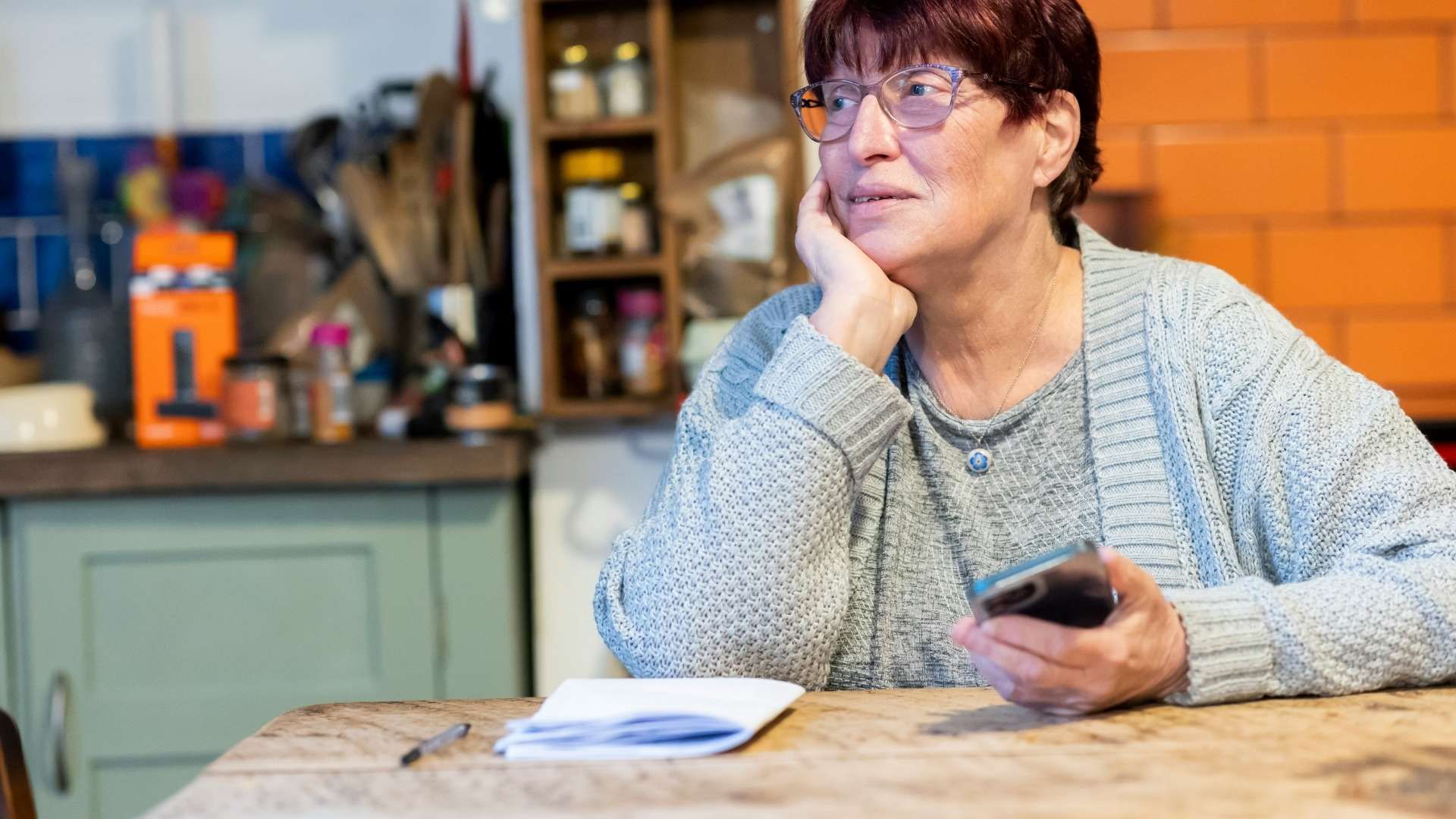 Person seated at a kitchen table holding a phone with papers and a notebook nearby