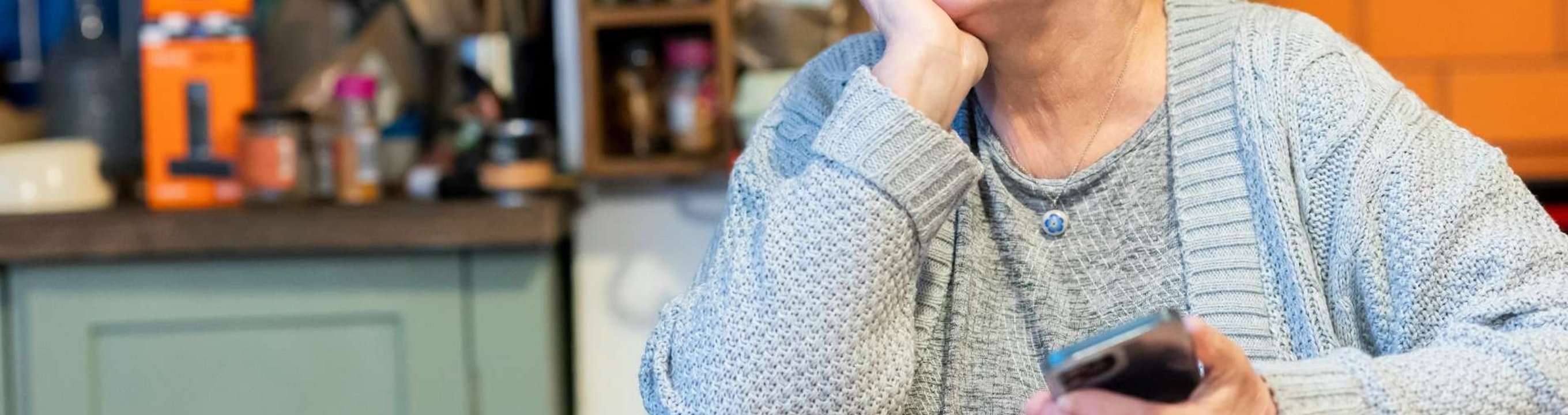 Person seated at a kitchen table holding a phone with papers and a notebook nearby