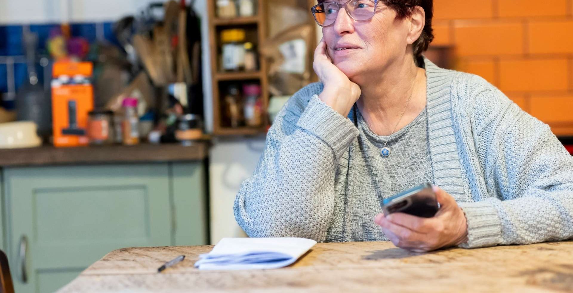 Person seated at a kitchen table holding a phone with papers and a notebook nearby