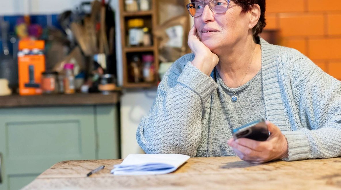 Person seated at a kitchen table holding a phone with papers and a notebook nearby