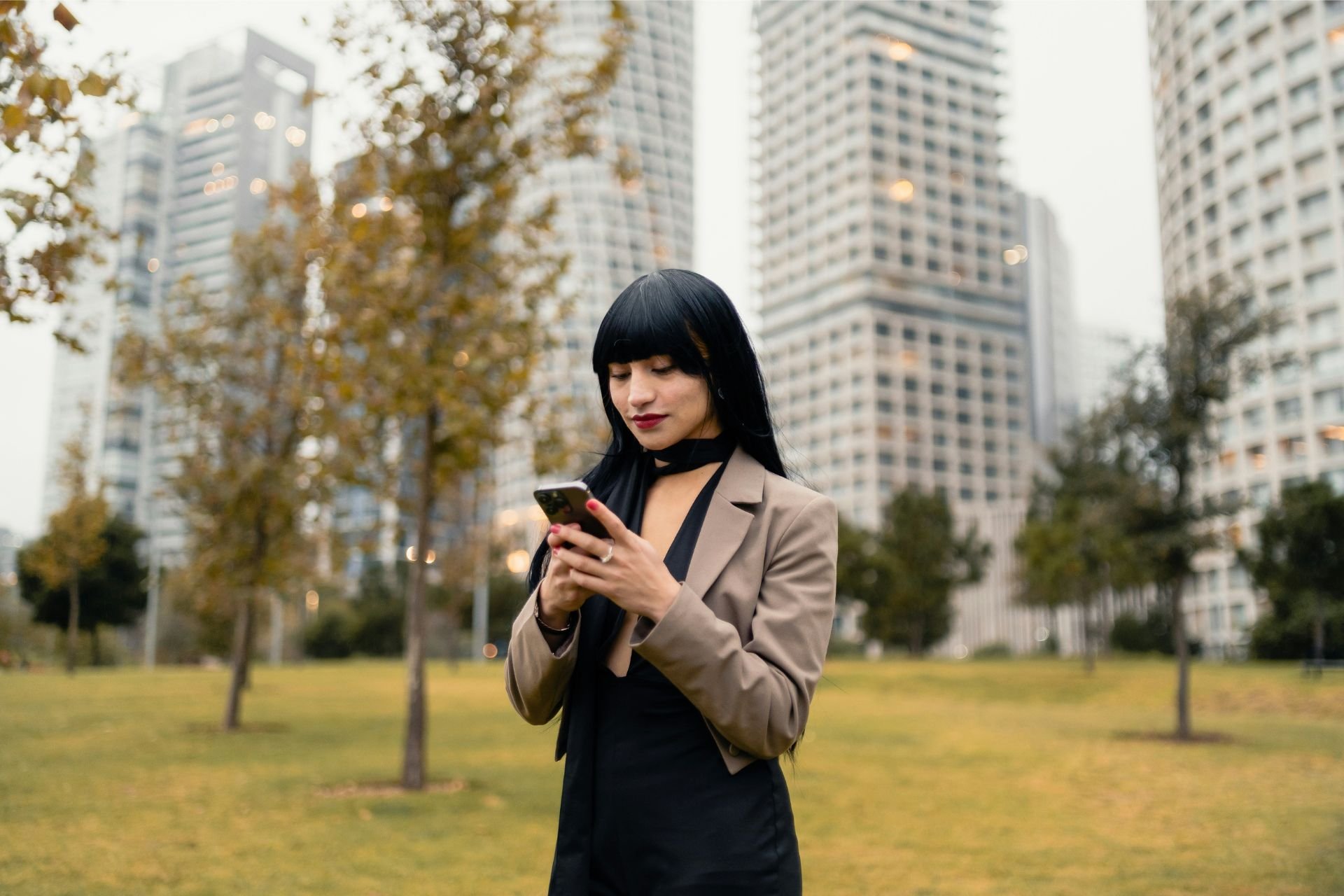 Person using a smartphone in a park with city buildings behind them.