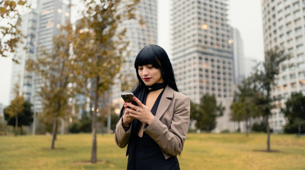 Person using a smartphone in a park with city buildings behind them.