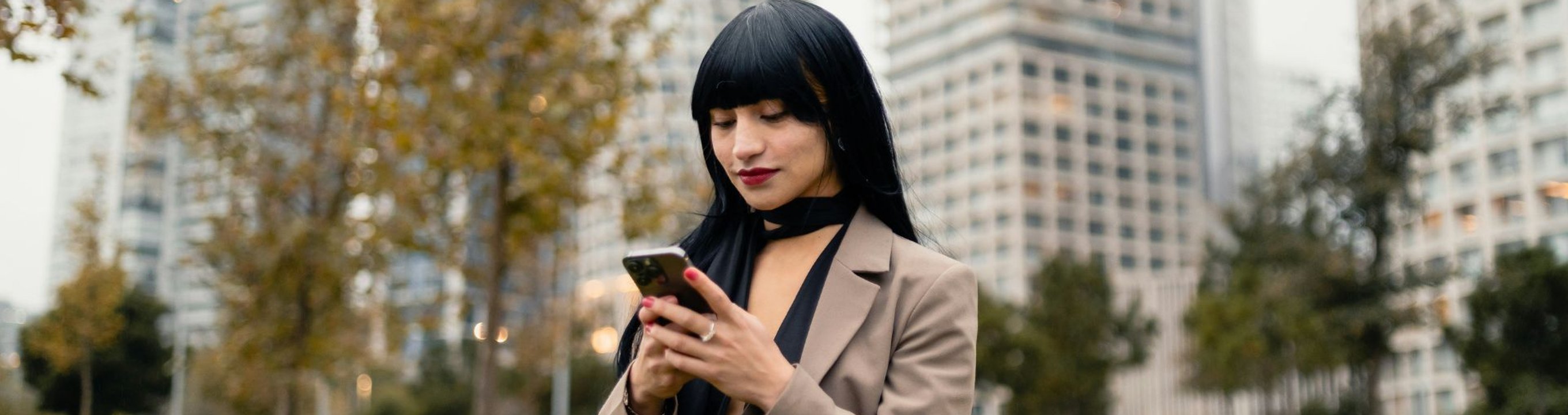 Person using a smartphone in a park with city buildings behind them.