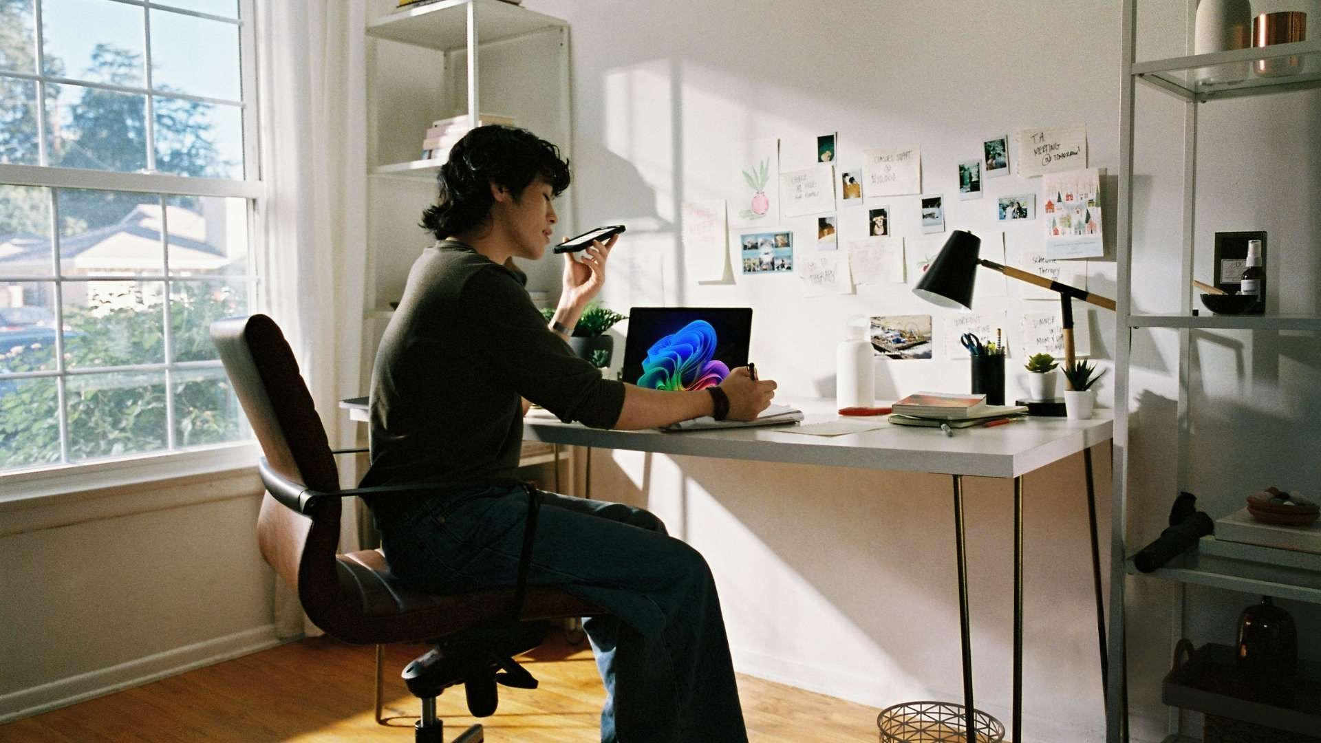 Person seated at a desk using a laptop while speaking into a smartphone