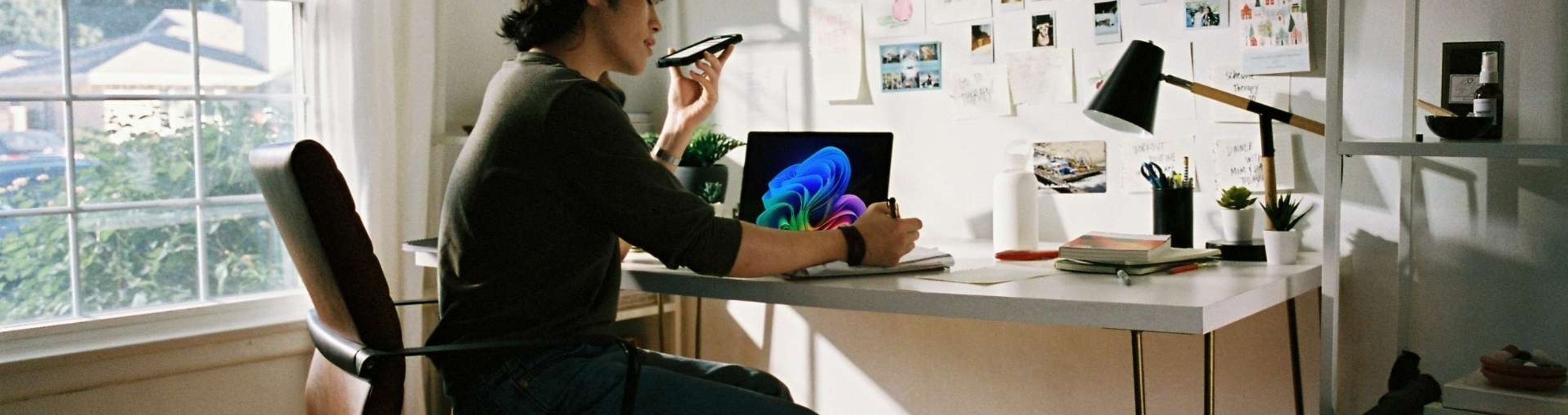 Person seated at a desk using a laptop while speaking into a smartphone