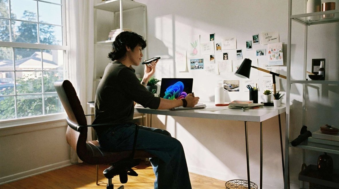 Person seated at a desk using a laptop while speaking into a smartphone