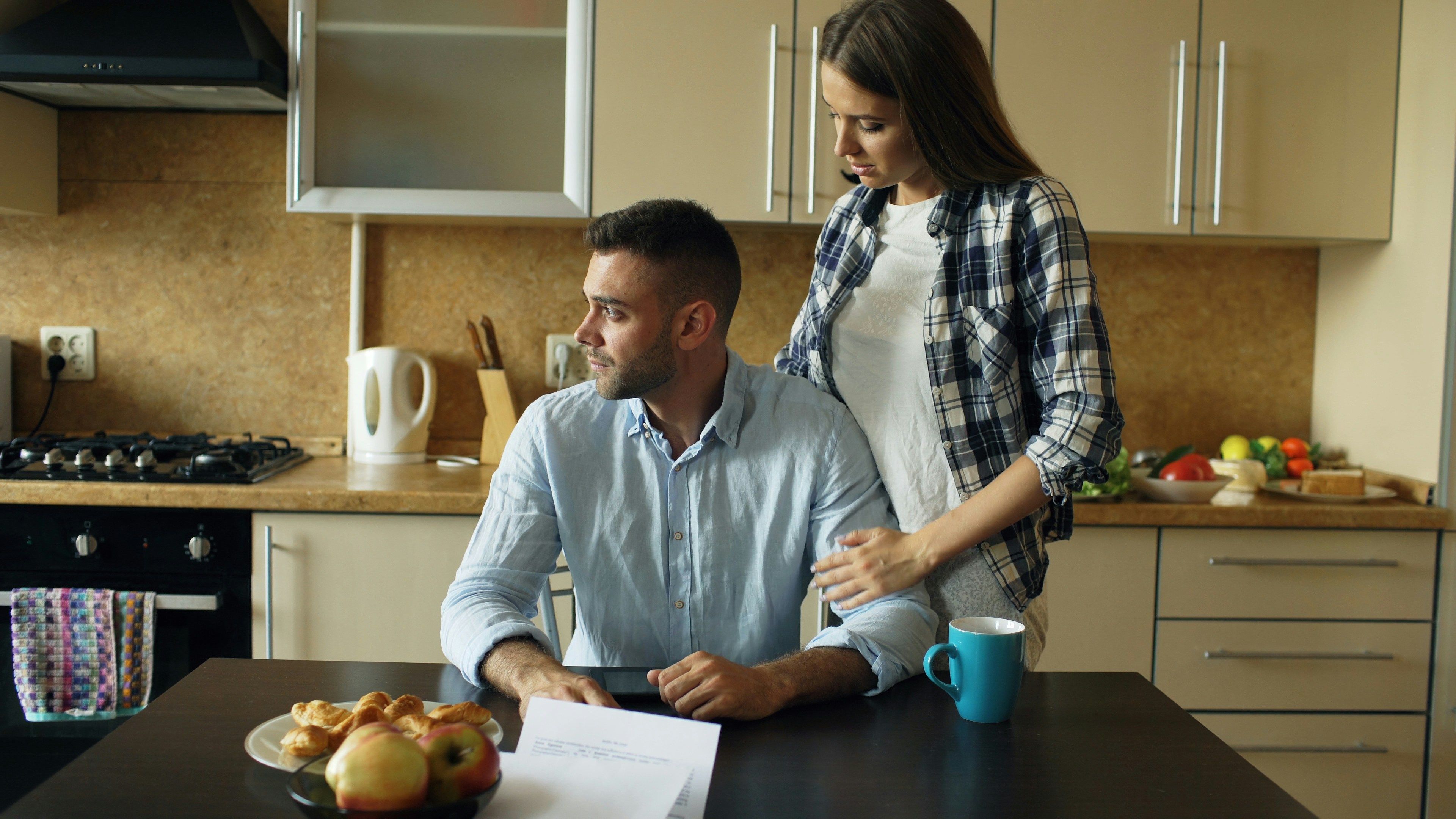 A couple sits at a kitchen table reviewing bills, looking concerned about their finances.