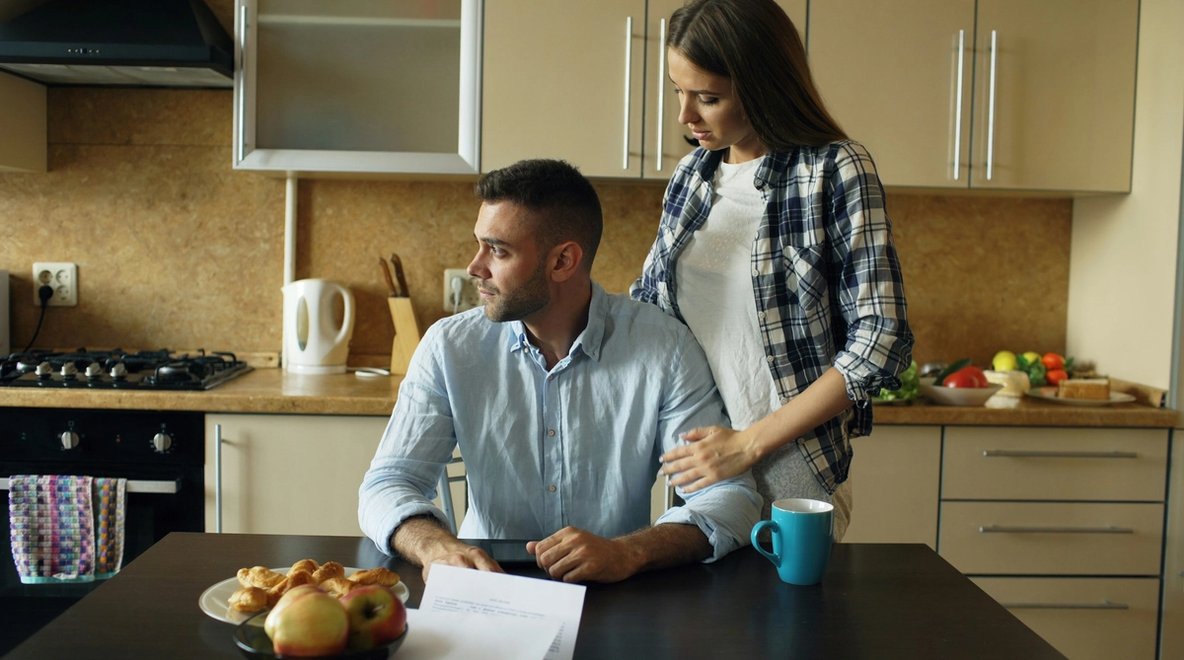 A couple sits at a kitchen table reviewing bills, looking concerned about their finances.