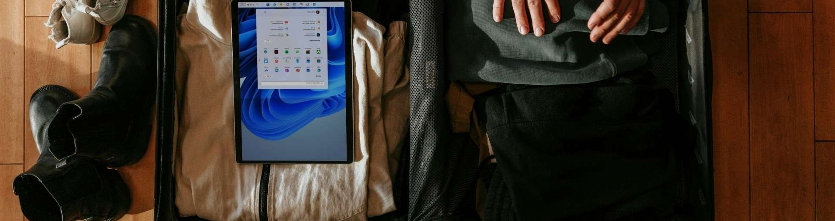 Open suitcase on a wooden floor with clothes, shoes and a tablet packed beside a bag