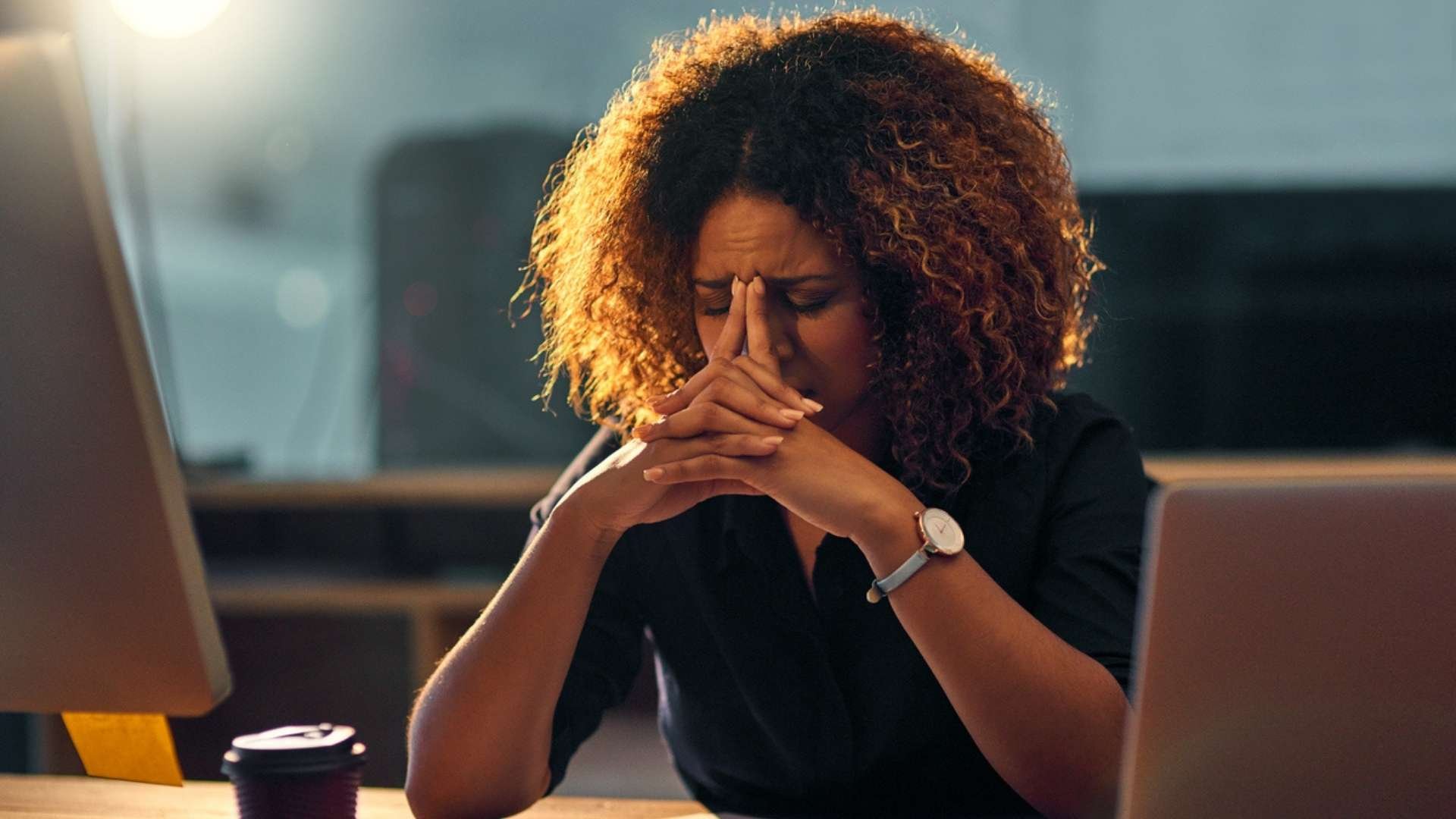 Person sitting at a desk with a laptop, hands clasped in front of their face.