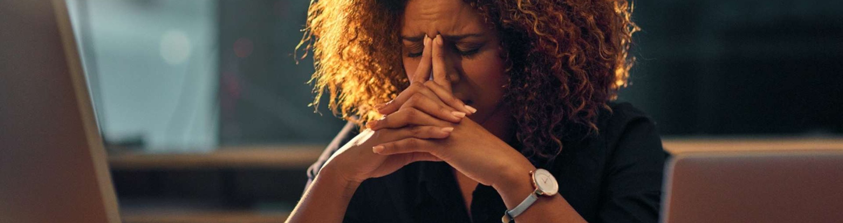 Person sitting at a desk with a laptop, hands clasped in front of their face.