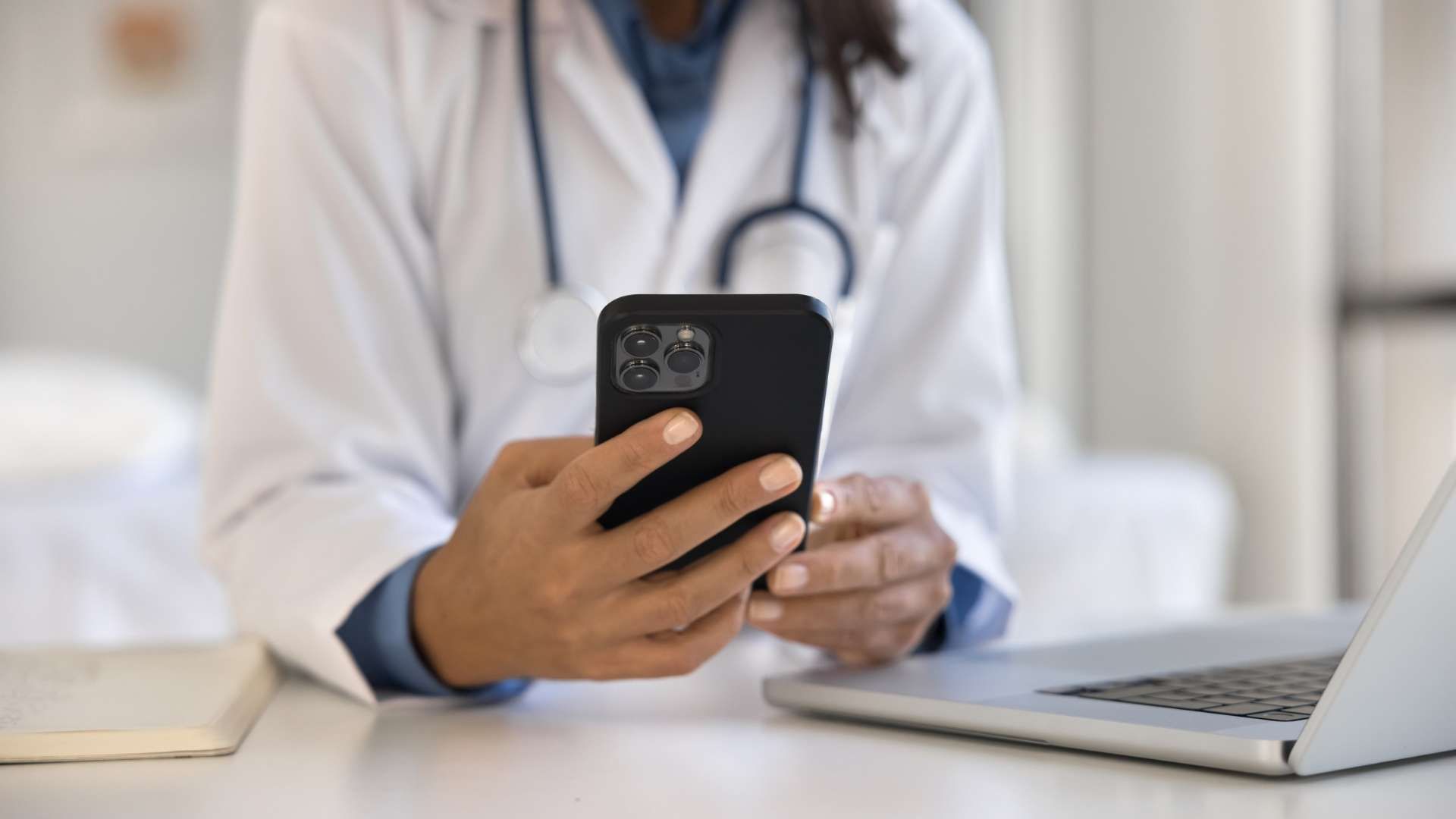 Person in a medical setting wearing a stethoscope using a smartphone beside a laptop