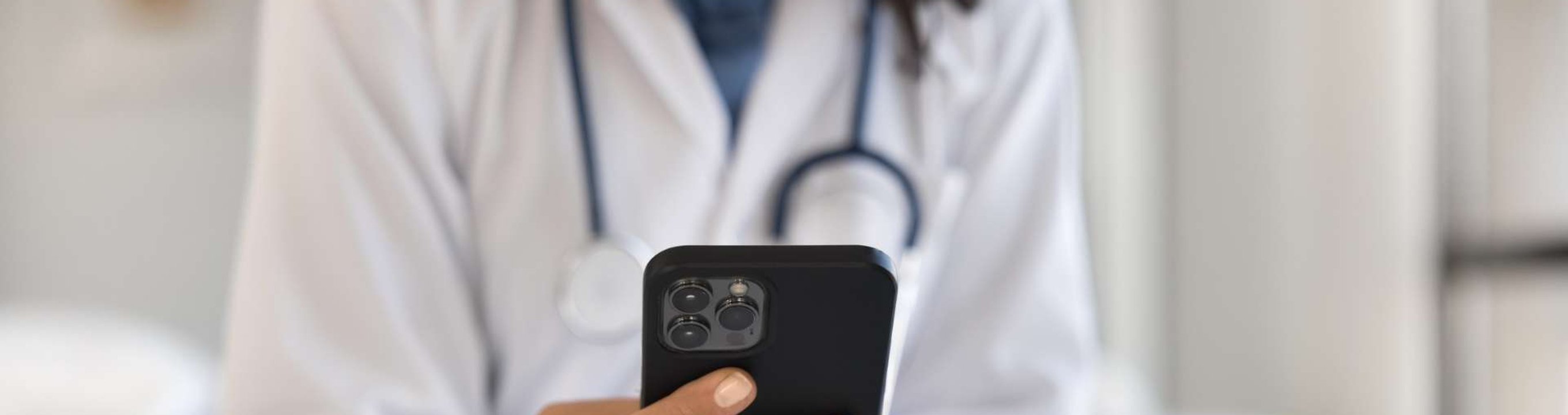 Person in a medical setting wearing a stethoscope using a smartphone beside a laptop