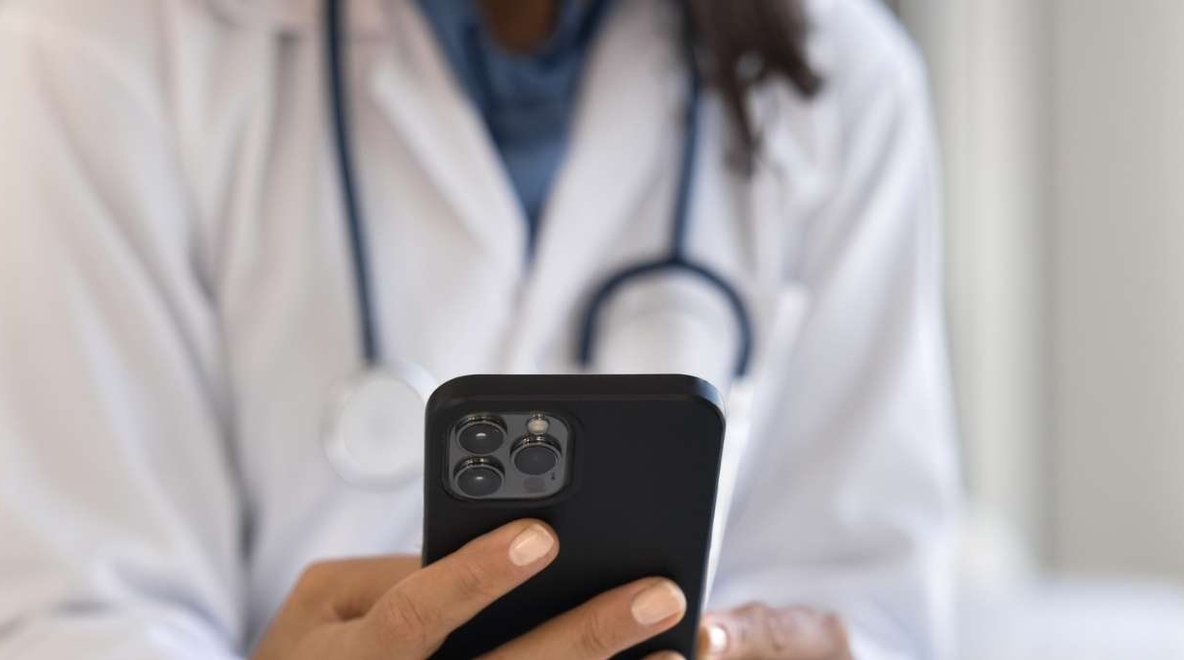 Person in a medical setting wearing a stethoscope using a smartphone beside a laptop