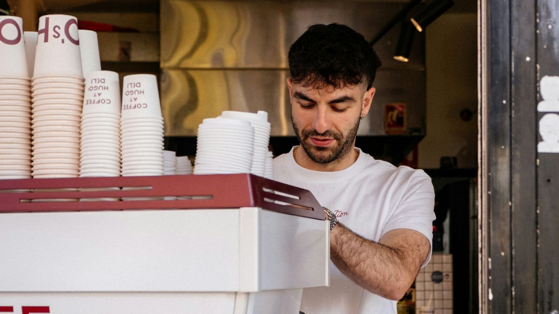 Person working behind a café counter with stacks of takeaway coffee cups and equipment visible.