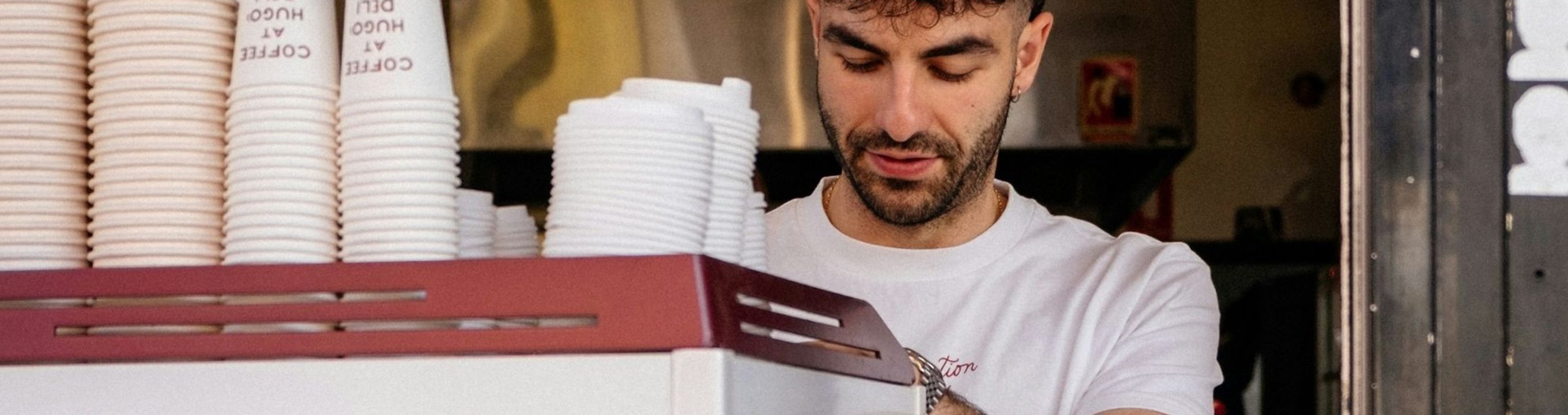 Person working behind a café counter with stacks of takeaway coffee cups and equipment visible.