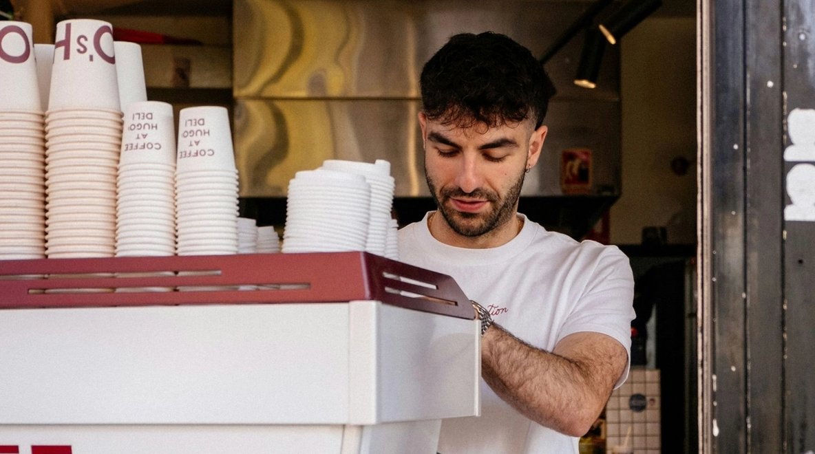 Person working behind a café counter with stacks of takeaway coffee cups and equipment visible.