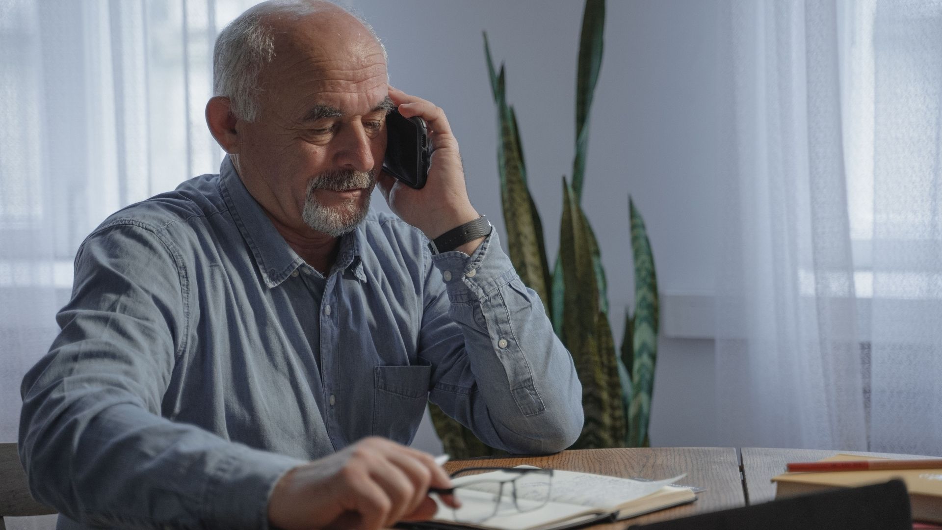 Older person talking on a mobile phone while seated at a table in a home setting.