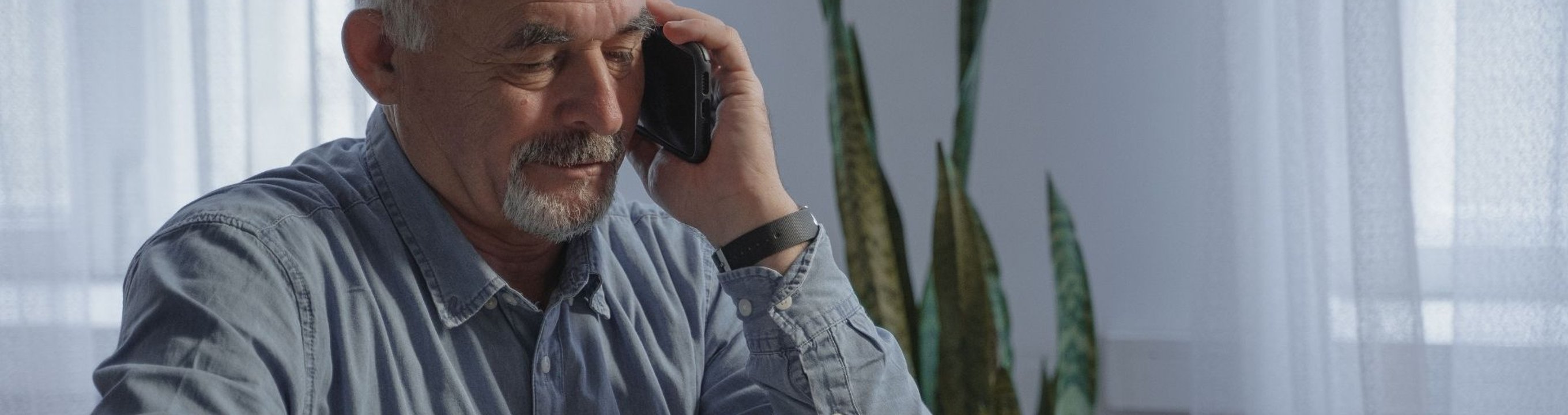 Older person talking on a mobile phone while seated at a table in a home setting.