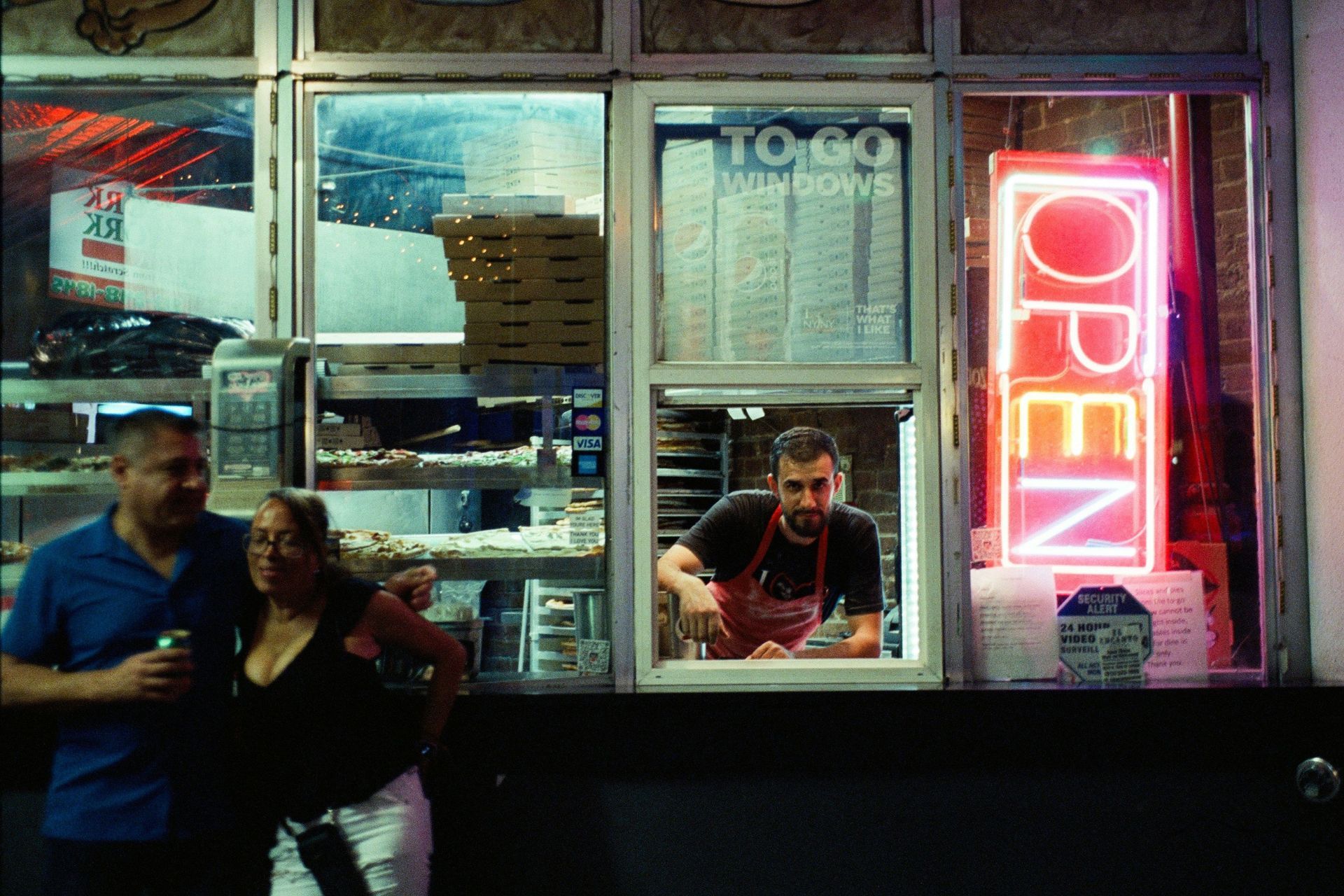 Man serving customers at a takeaway shop window with a neon “open” sign.