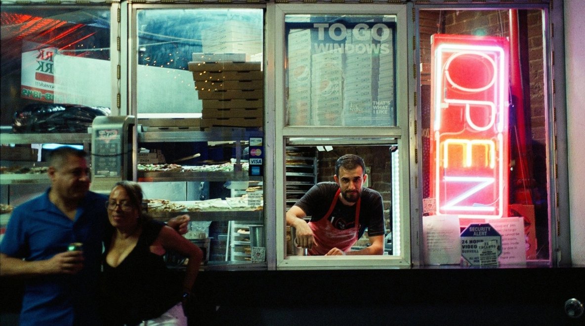 Man serving customers at a takeaway shop window with a neon “open” sign.