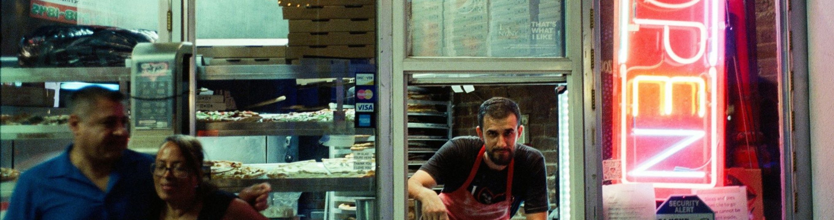 Man serving customers at a takeaway shop window with a neon “open” sign.