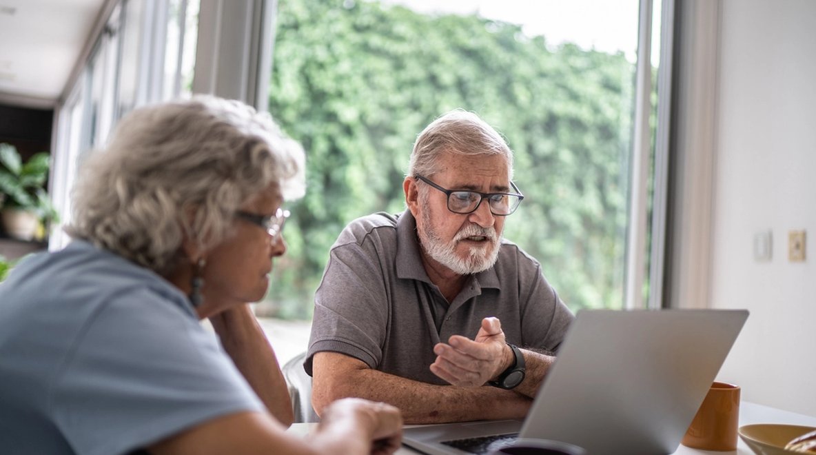 Two people sitting at a table looking at a laptop together near a window.