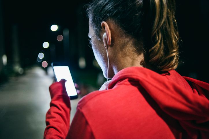 Person in a red hoodie using a smartphone outdoors at night with earphones and street lights in the background