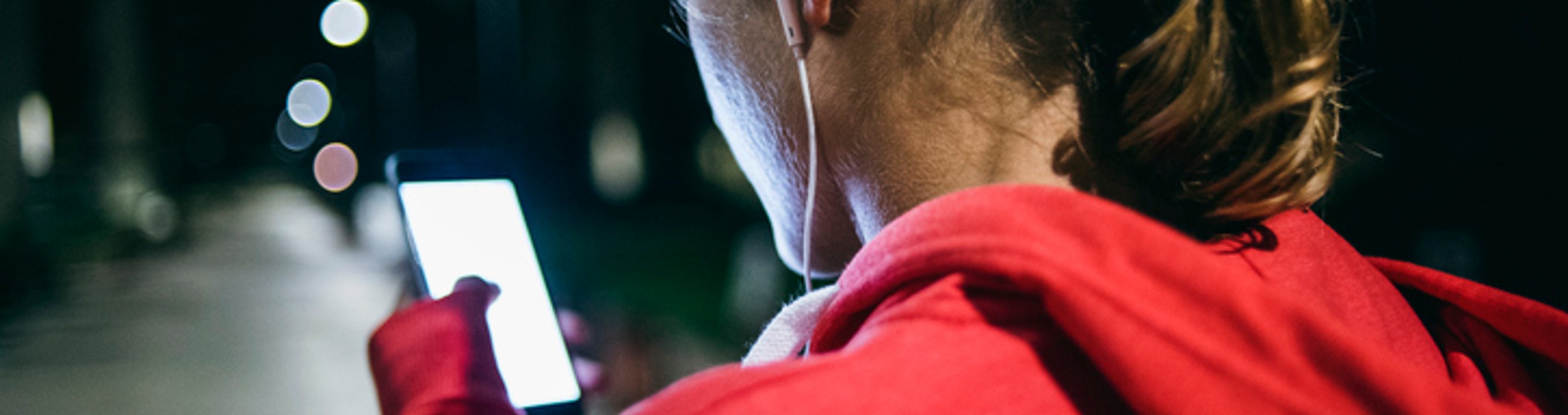 Person in a red hoodie using a smartphone outdoors at night with earphones and street lights in the background