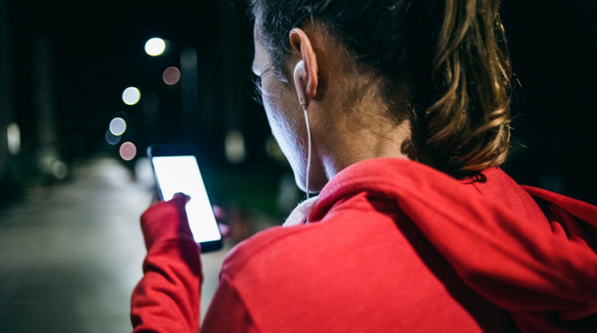 Person in a red hoodie using a smartphone outdoors at night with earphones and street lights in the background