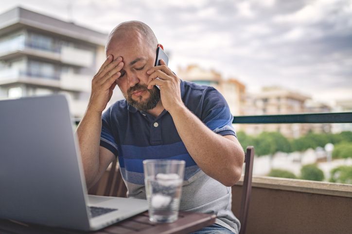 Person sitting at an outdoor table using a laptop while holding a phone to their ear