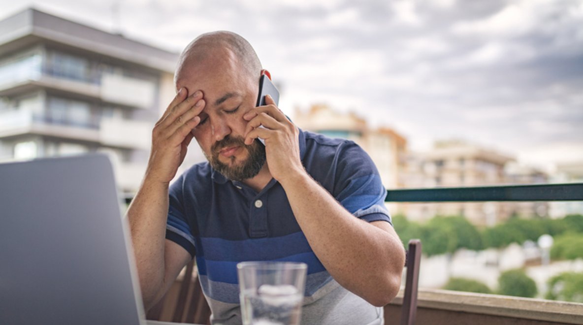 Person sitting at an outdoor table using a laptop while holding a phone to their ear