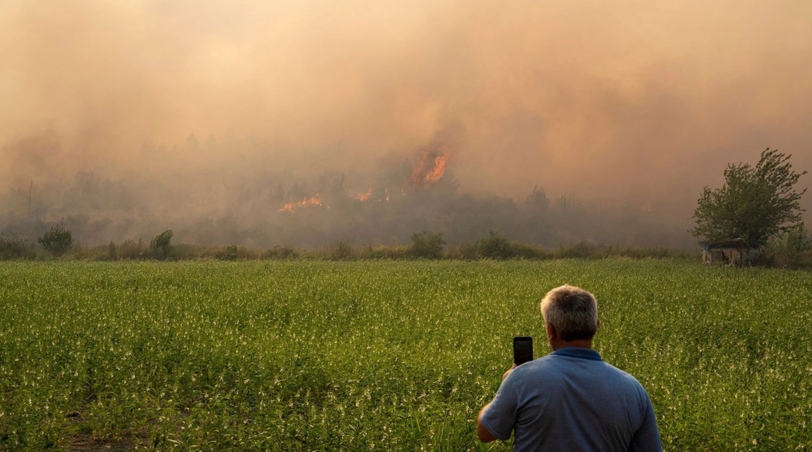 Person holding a mobile phone while watching smoke and flames from a bushfire in the distance.